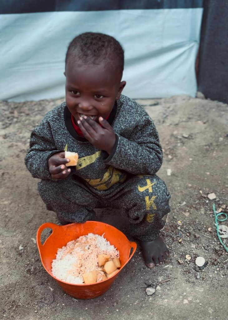 Kind op hurken eet uit oranje bak met rijst tijdens voedselhulp, buiten op zandgrond.