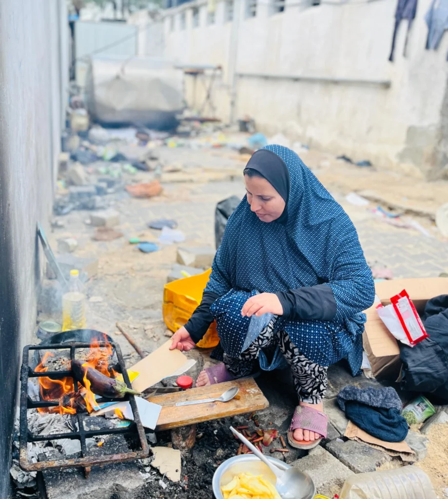Vrouw in een verwoeste straat die op een geïmproviseerd vuurtje eten bereidt tussen puin en schaarse spullen, symbool voor het dagelijks overleven in Gaza.
