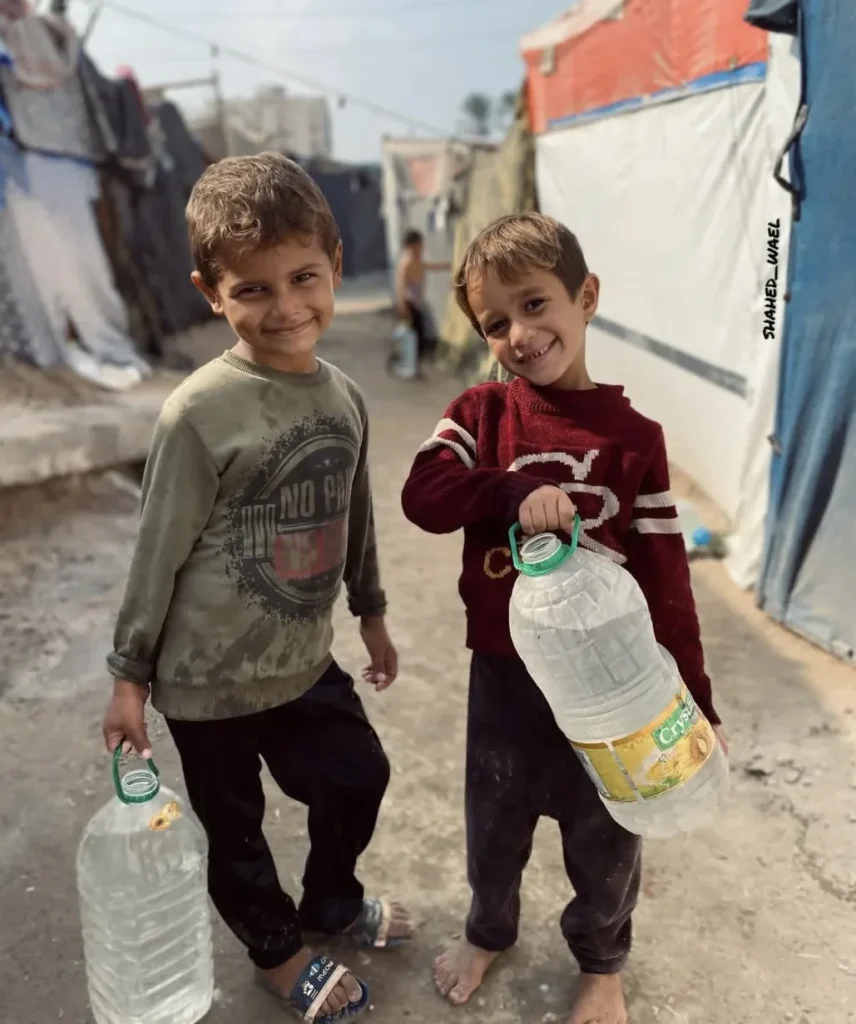 Twee kinderen dragen grote waterflessen in een kamp, symbool voor toegang tot schoon drinkwater