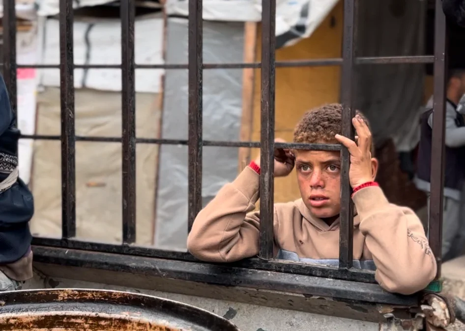 Jongen achter hek kijkt verlangend naar pan met warme maaltijd tijdens voedseluitdeling in Palestina.