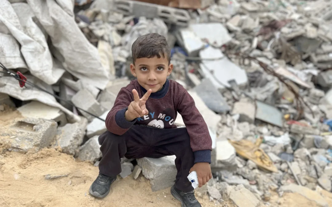 Jongen zit op puin in Gaza en maakt vredesteken met zijn hand, voor ingestorte gebouwen op de achtergrond