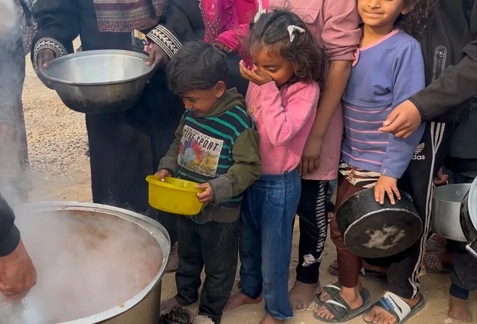 Kinderen in de rij voor voedsel bij een vluchtelingenkamp met dampende pan op de voorgrond