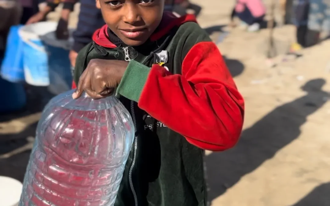 Jonge jongen in vluchtelingengebied tilt een grote jerrycan met water terwijl anderen op de achtergrond wachten