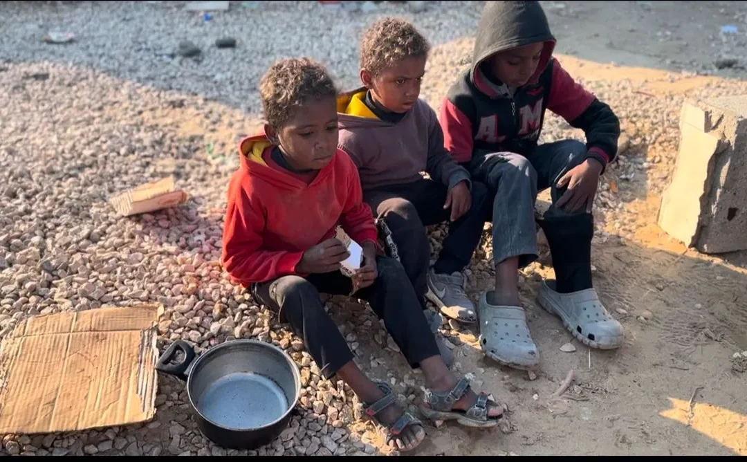 Drie jonge kinderen zitten op de grond met lege pannen en karton bij zich, wachtend op voedsel in een kampomgeving