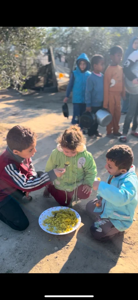 Kinderen delen een bord met rijst op de grond terwijl anderen in de rij wachten met lege pannen