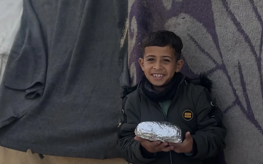 Smiling young boy holding a foil-wrapped meal in front of a tent wall.