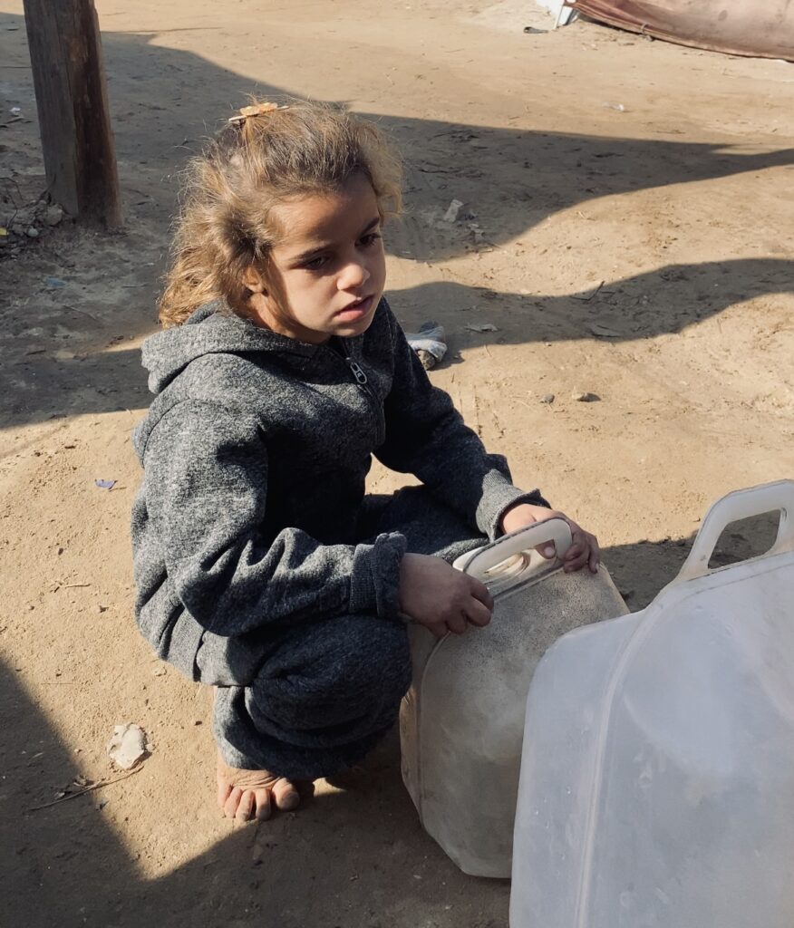 Jong meisje in Gaza zit op de grond met een lege jerrycan in haar handen, wachtend op schoon water dat door Stichting ULFAH wordt geleverd.