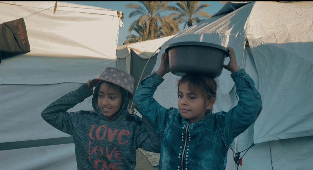 Twee kinderen in een tentenkamp, waarvan één een teil op het hoofd draagt, tussen witte tenten en palmbomen.
