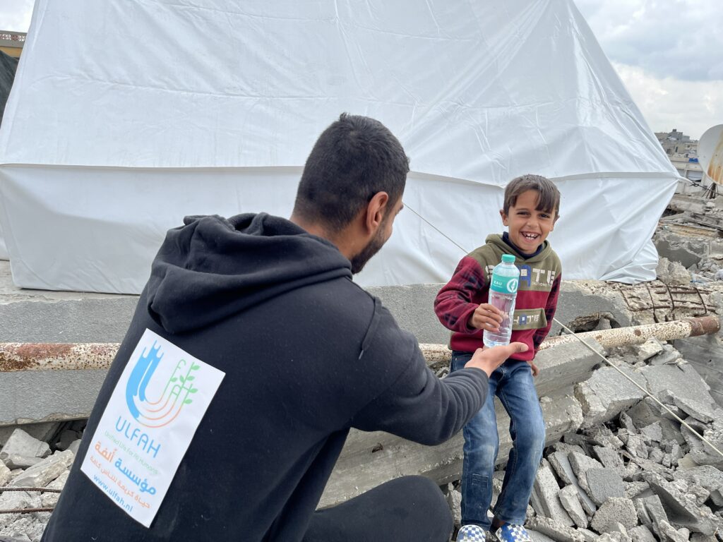 Vrijwilliger van Stichting ULFAH geeft een lachend kind een fles water tussen puin en noodtenten.