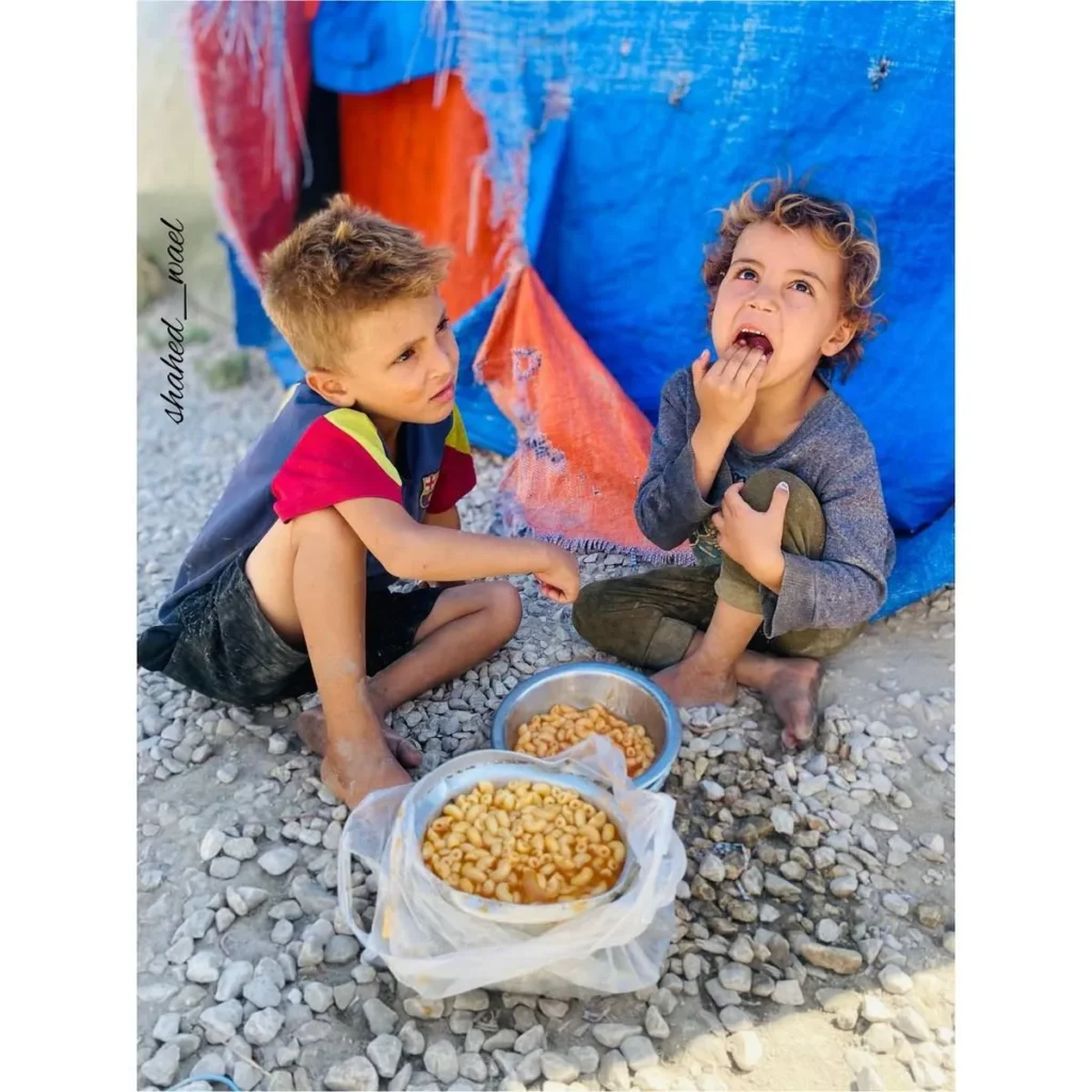 Twee jonge kinderen die buiten op de grond zitten en een kom warme pasta delen in een noodsituatie.