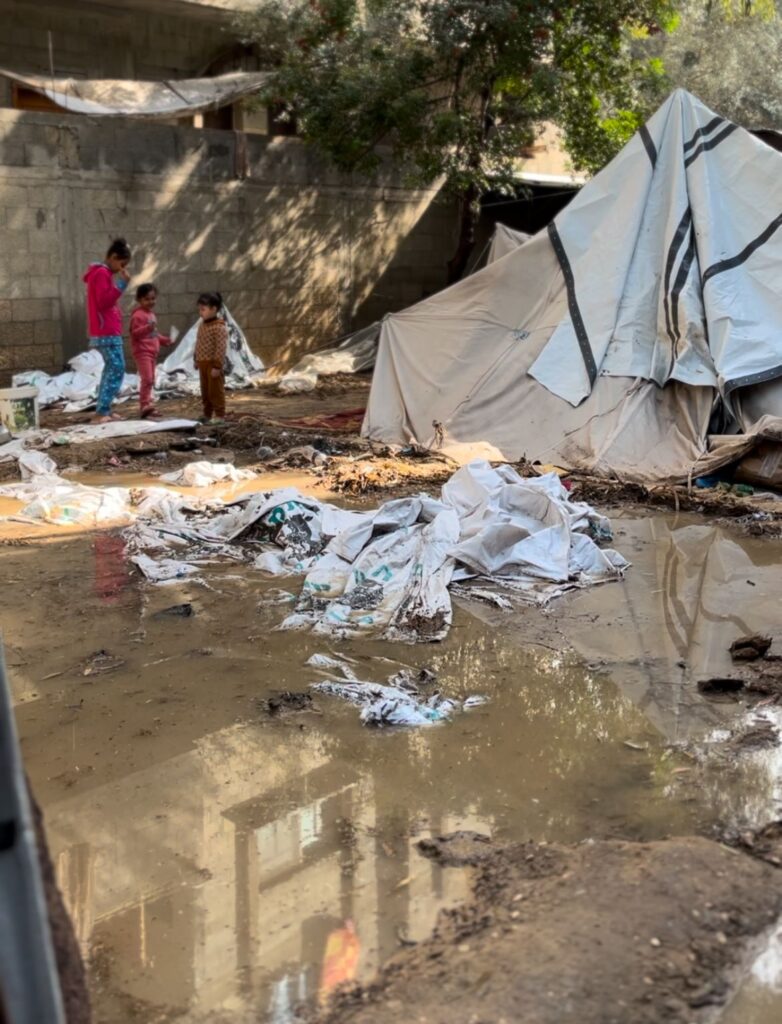 Drie kinderen naast een noodtent, omringd door modderplassen en natte zeilen in een geïmproviseerd kamp