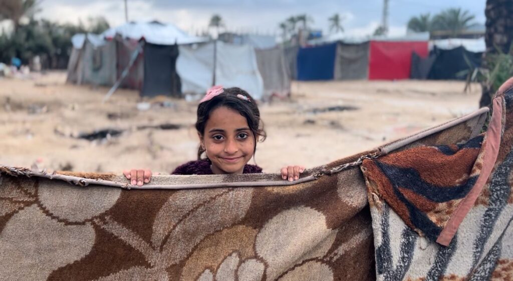 Palestijns meisje glimlacht achter een versleten deken in een geïmproviseerd tentenkamp.