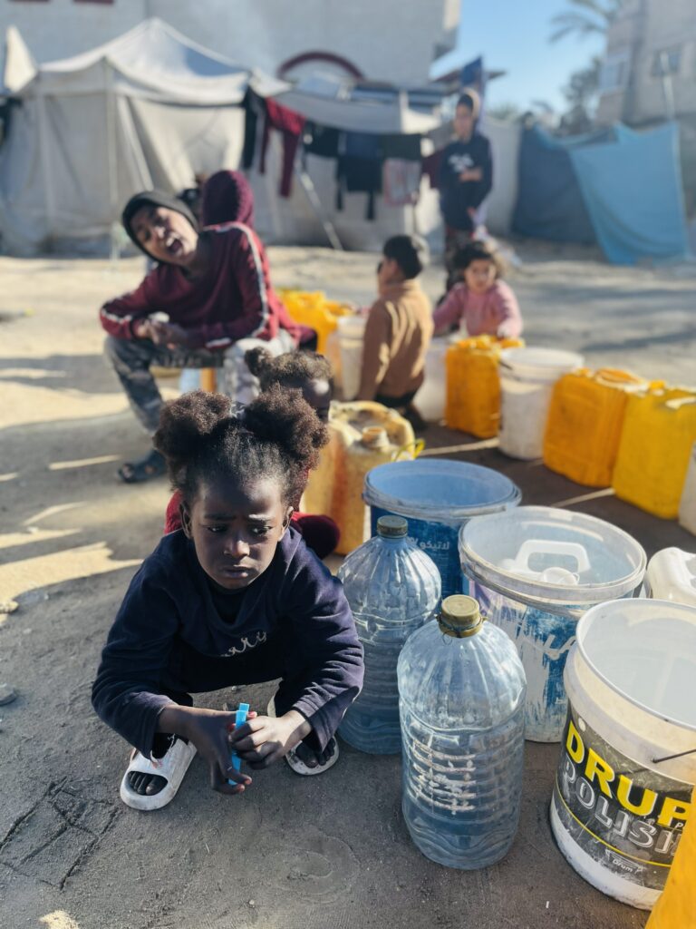Kinderen in een tentenkamp met jerrycans en waterflessen, wachtend op schoon drinkwater.