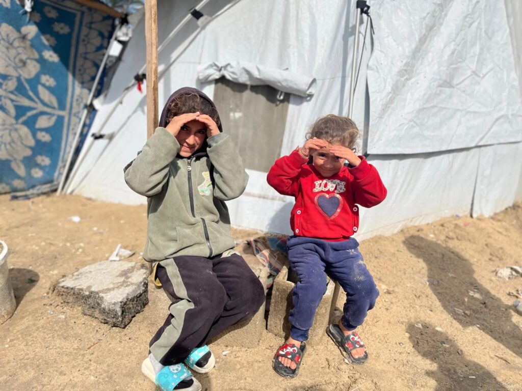 Twee jonge kinderen zittend voor een tent in een vluchtelingengebied, in de zon met hun hand boven de ogen.